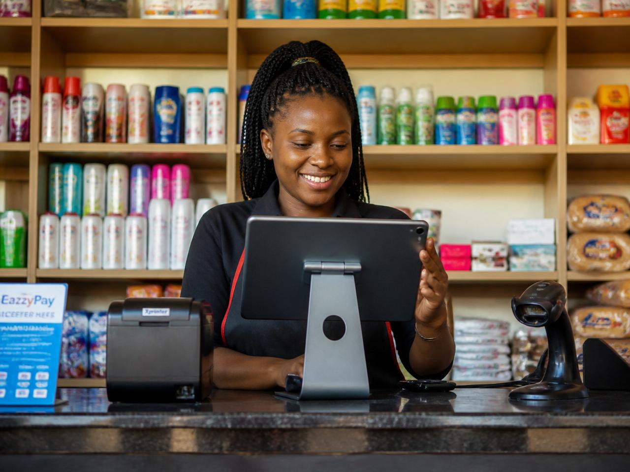 Shop attendant in a Kenyan minimart using the Zeni POS point of sale system on a tablet with a receipt printer and barcode scanner